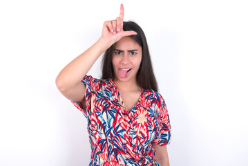 young beautiful brunette woman wearing colourful dress over white wall gestures with finger on forehead makes loser gesture makes fun of people shows tongue
