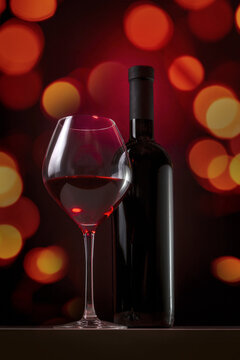 A bottle and a glass of red wine on a dark background with bright lights out of focus. Festive image of wine