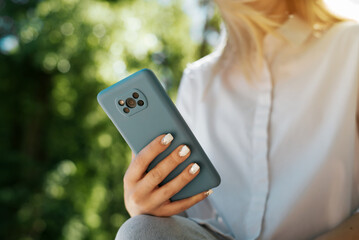 Unrecognizable woman in white shirt using smartphone outdoors. Close-up of female caucasian hands holding mobile phone on sunny summer day in park. Low angle view, selective focus on gadget, bokeh