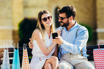 Portrait of beautiful smiling young couple sitting on a bench after shopping using phone.