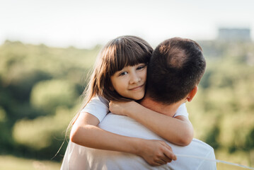 Young girl hugs her father, putting arms around his neck and looking at the camera. Outdoor family walk