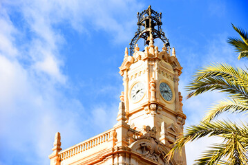 Fototapeta na wymiar Clock tower building. Large clock tower with hour hands that shows the time of day and night. Clock tower against the background of palm trees and blue sky. Clock tower. Clocks with time on building.