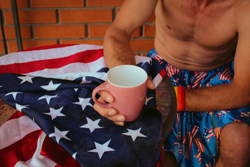 Closeup pink cup in hands of shirtless LGBT+ man, with Americam flag on the table. Symbol of...