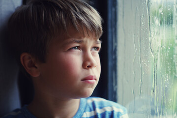 Bored of being bored. Shot of a sad young boy watching the rain through a window at home.