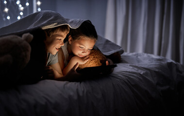 They stay entertained with technology. Shot of two adorable little siblings using a digital tablet together in bed at night.