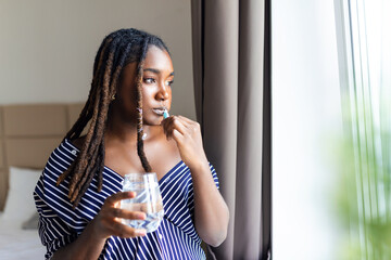 Young African woman brushing teeth with toothbrush, holding glass of water and looking in window