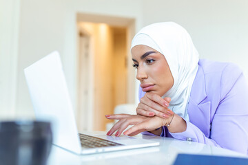 Fototapeta na wymiar Frustrated young muslim businesswoman wearing hijab working on a laptop sitting at desk in office.