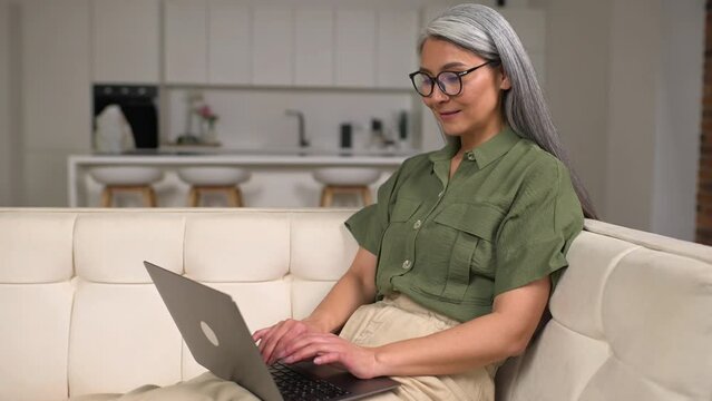 Senior asian female businesswoman or freelancer in comfy casual clothes sitting on the couch and working on the project, looking at the screen with pleasure smile