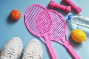 Summer sport activities flat lay object photography. White sneakers, pink rackets, dumbbells, mandarin, lemon and clean water on a blue background. Flat lay fitness image
