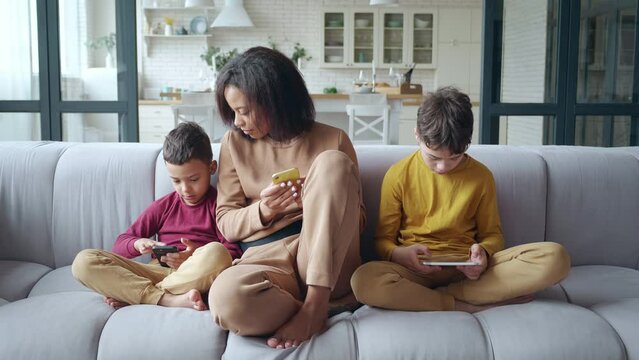 A pretty young African American woman surfing her smartphone turns to her young son actively swiping his cell phone. Boy having fun sitting on sofa with digital tablet next to his family. Day off