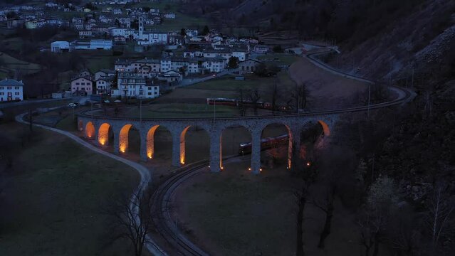 Train on Illuminated Brusio Spiral Viaduct in Switzerland in the Evening. Bernina Railway. Swiss Alps. Aerial View. Drone Flies Backwards and Upwards