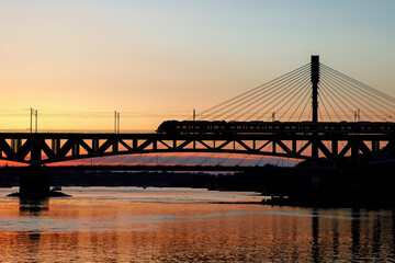 The train riding over the bridge over the river at sunset.