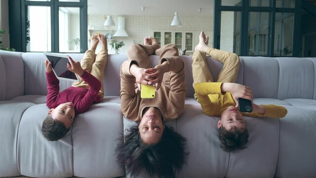 Young mother and children on the couch with their feet up, everyone is busy with their phone. African American family resting on the couch on day off. Woman with sons having fun using digital gadgets