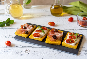 Grilled polenta with stracchino cheese, tomatoes, herbs, speck and olive. White wooden background, ingredients and table wine.