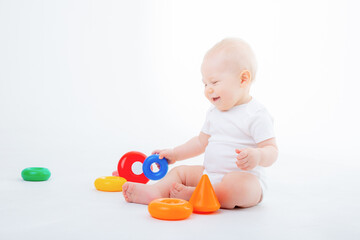 a baby boy in a white bodysuit is sitting playing with a multicolored pyramid smiling on a white background