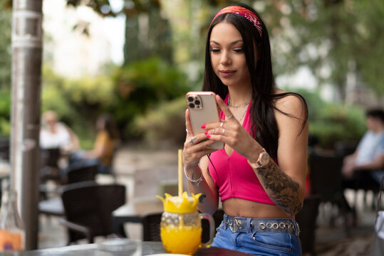 Woman enjoys an orange drink on the terrace of a bar while browsing the internet through her mobile phone