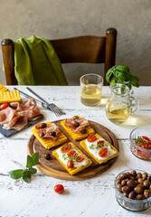 Bruschettas with grilled polenta topped with stracchino cheese, tomatoes, herbs, speck and olives. Chair, napkin in the back.
