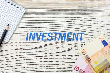 Naklejka premium INVESTMENT - word (text) and euro money on a white wooden table, notebook, notepad. Business concept: buying, selling, commerce (copy space).
