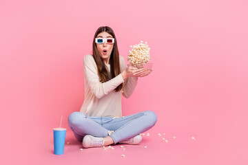 Obraz na płótnie Canvas Portrait of attractive amazed girl sitting on floor watching video throwing corn isolated over pink pastel color background