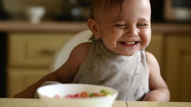 A cute one-year-old baby sits at home in the kitchen on a high chair and eats rye flakes, the child eats a dry breakfast and laughs sweetly