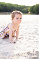 Fototapeta na wymiar happy baby girl sitting on the beach on sand wearing striped summer dress on sunny day