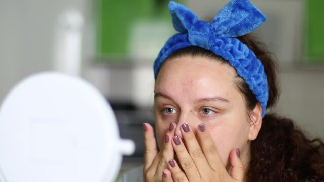 Young curly woman in blue hairband use and rubbing hydrating moisturizing cream or lotion, doing stretching skin patting massage. Skin vitamin absorption for healthcare, anti-aging detox effect
