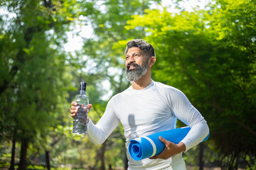 Mature beard indian fitness man wearing white sportswear walking with water bottle and yoga mat roll in hand at park.