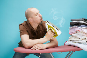A funny bald man with emotions using electrical iron on ironing board over on blue background.