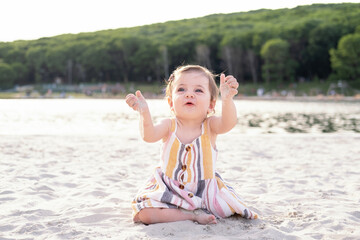 happy baby girl sitting on the beach on sand wearing striped summer dress on sunny day