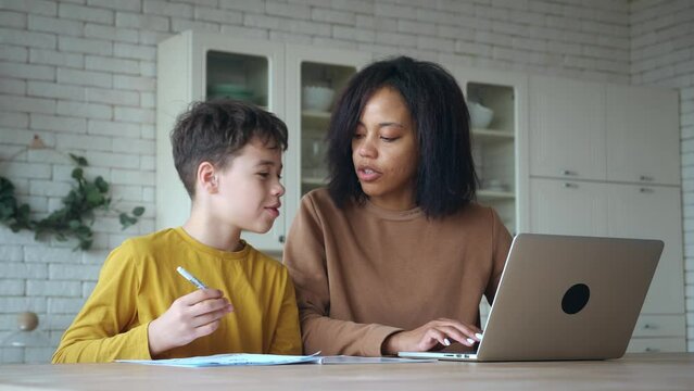 Patient mom stares at her son, an elementary school student, reluctant to do his homework as she explains his lessons. Younger brother crawls under the table, interfering with lessons. Online study