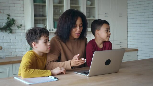 African American woman and sons sitting at the table in the kitchen ,waving hands looking at web camera using laptop for video call. Smiling mom and children boys greeting online making video call