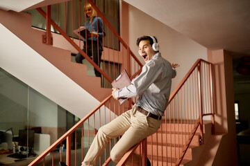 Playful businessman with headphones sliding down the railing in hallway of an office building.