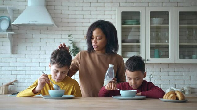 Cute boys sitting at the table for breakfast with chocolate flakes. Charming African-American mom standing behind them with a bottle of milk and persuading the eldest son, playing with a spoon to eat