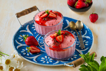 Dessert, airy strawberry mousse in glass bowls, decorated with fresh strawberries, syrup and chocolate chips on a blue tray on a light concrete background.