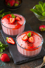 Dessert, airy strawberry mousse in glass bowls, garnished with fresh strawberries, syrup and chocolate chips on a black slate board against a dark concrete background.