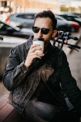 Fototapeta na wymiar young man in sunglasses in the summer sits on a city street and drinks coffee. hipster man