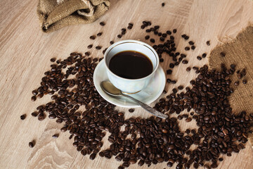 A cup of hot coffee in a plate with a spoon. Coffee beans are scattered on a wooden light brown table.