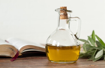 Pure olive oil in a glass jar, an open Bible Book, and a green olive branch on a wooden table with...