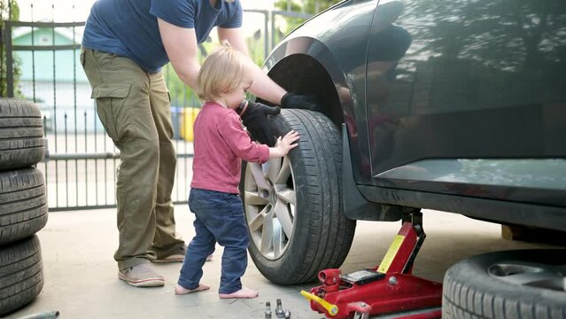 Cute toddler boy helping his father to change car wheels at their backyard. Father teaching his little son to use tools. Active parent of a small child.