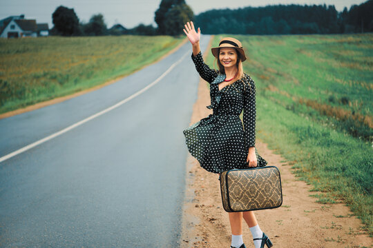 happy woman in a hat travels with a suitcase on the road hitchhiking and waving hand. retro style.