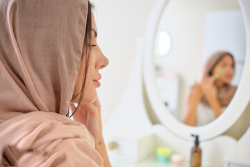 young Muslim woman wearing hijab headscarf putting on makeup. Muslim woman applying make-up against blue colour background. Young Muslim woman with mirror and makeup brush and jade
 - obrazy, fototapety, plakaty