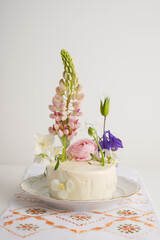 Tiny wedding cake with fresh flowers with lupine, on a light background with local confectionery. Vegan wedding white cake on light background