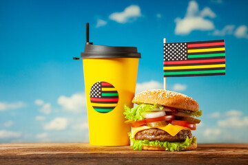 Classic American burger with alternative African American juneteenth flag on the top and black paper cup with straw over blue sky background. Close-up with selective focus.