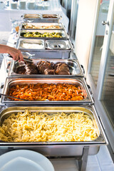 Rich, buffet style, self-serving food at a wedding party. Hand reaching out to grab some meat to put on the plate. Vegetables meat, fish, rice, and noodles. 