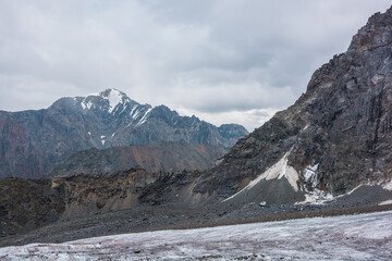 Awesome landscape with long glacier tongue against high mountain range with sharp rocky peaked top in overcast weather. Cone shaped pointy peak in dramatic cloudy sky. Large glacier among sharp rocks.
