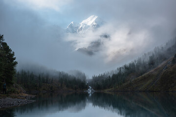 Tranquil scenery with snow castle in clouds. Mountain creek flows from forest hills into glacial lake. Snowy mountains in fog clearance. Small river and coniferous trees reflected in calm alpine lake.