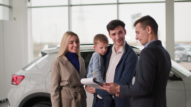 family in auto show, happy parents with male child talking to car salesman about new automobile model in showroom
