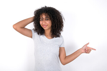 Surprised Young beautiful girl with afro hairstyle wearing grey t-shirt over white wall  pointing at empty space holding hand on head