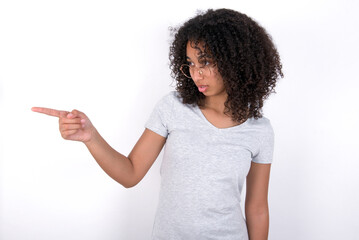 Stunned Young beautiful girl with afro hairstyle wearing gray t-shirt over white wall with greatly surprised expression points away on copy space, indicates something