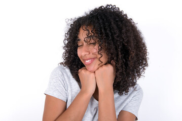 Cheerful Young beautiful girl with afro hairstyle wearing gray t-shirt over white background has shy satisfied expression, smiles broadly, shows white teeth, People emotions